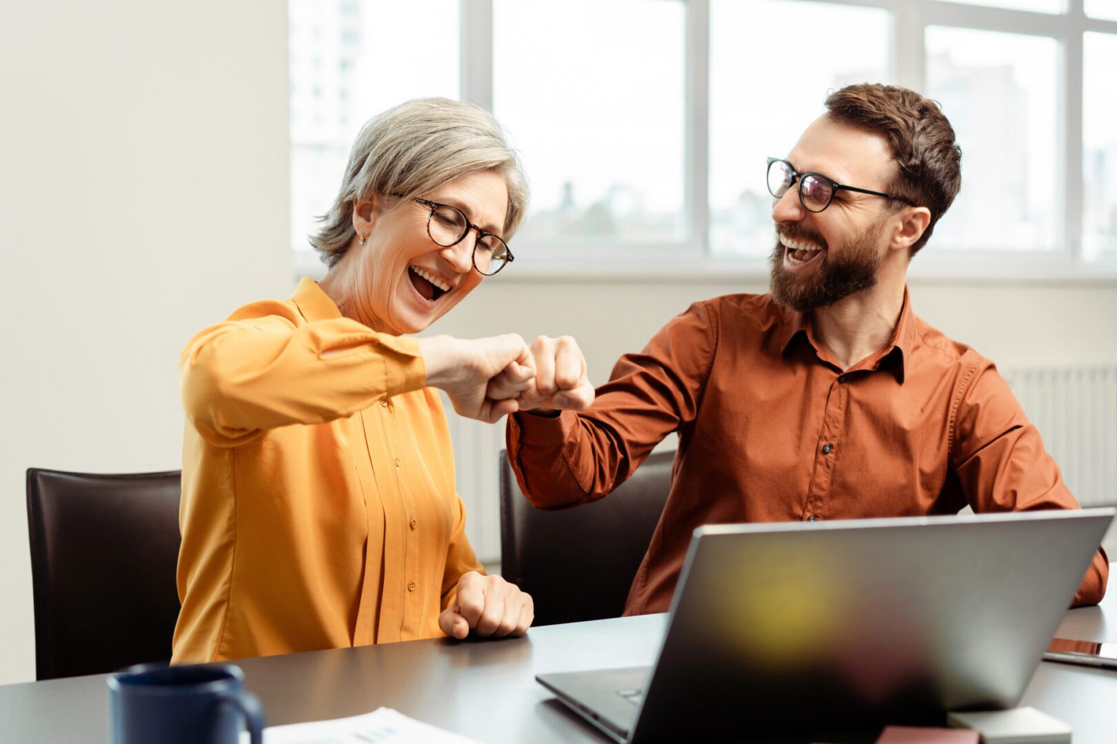 Overjoyed business colleagues bumping fists celebration success while working together. Happy senior woman and bearded man at modern office. Successful business concept